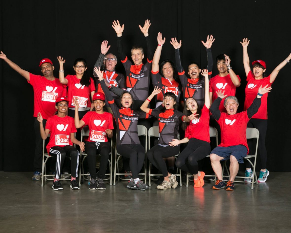 Becel Heart&Stroke Ride for Heart, large group of 15 people smiling and raising their hands up, a mix of wearing red t-shirts and a black and red biking uniform, black background